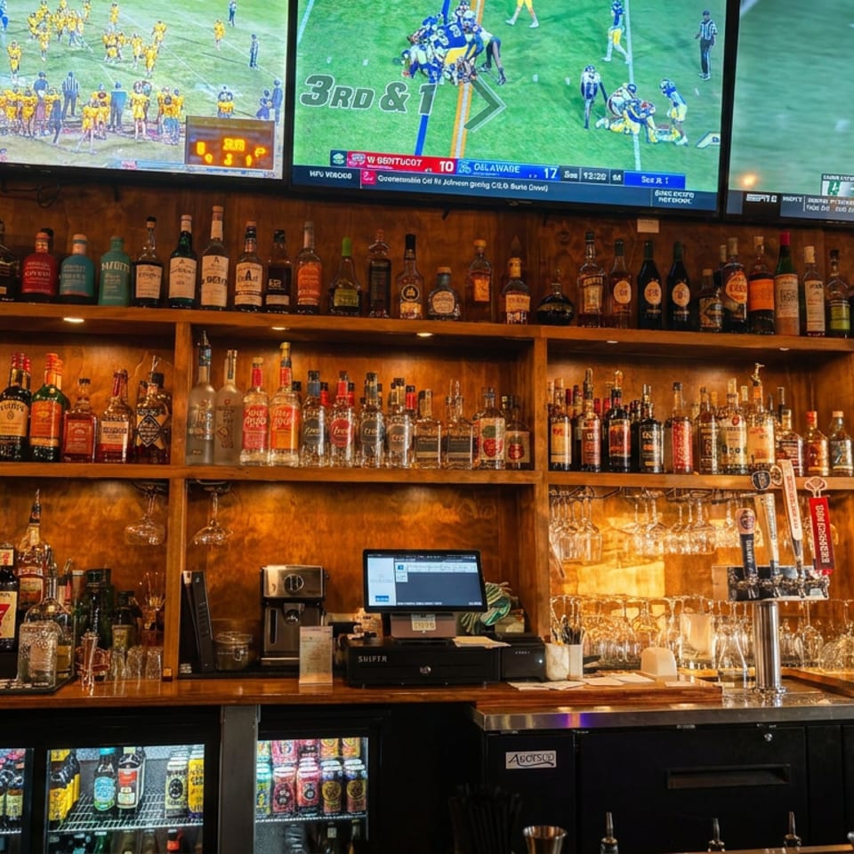 Warmly lit restaurant bar stocked with liquor bottles, glassware, taps, and televisions showing football games.