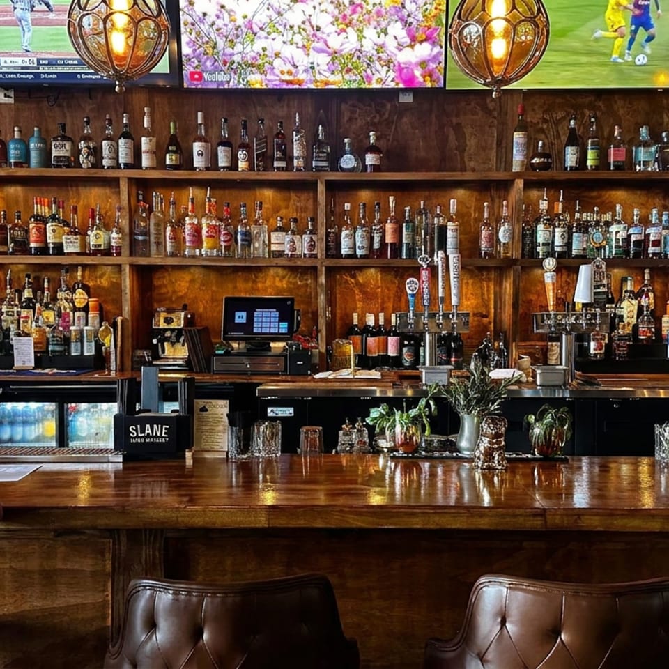 Polished wooden bar lined with liquor bottles, beer taps, sports screens, glowing pendant lights, and a seated patron.