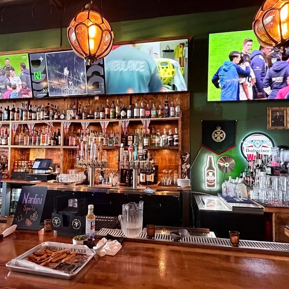 Sports bar interior with glowing pendant lights, televisions showing rugby, a liquor-filled wooden bar, beer taps, and fries on the counter.